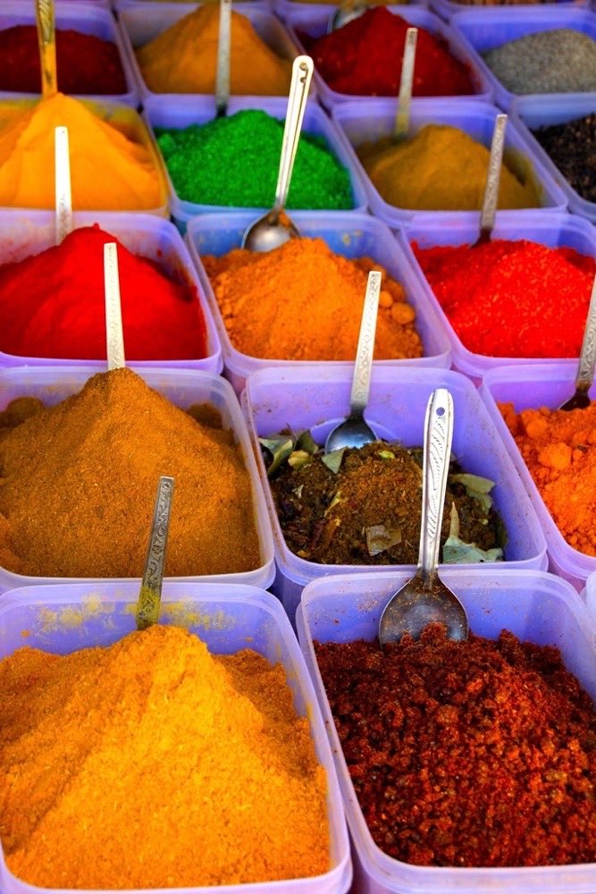 Market trays heaped with saffron, chilli, green and crimson spices, a spoon standing in each